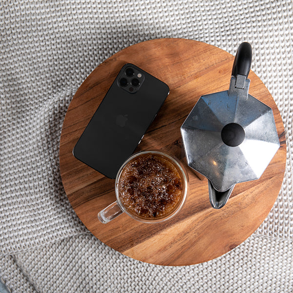 Wooden tray with a cup of coffee, phone, and metal coffee pot placed on an off white textured organic cotton throw.