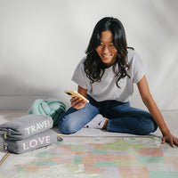 Woman sitting on a rug with two light grey travel blanket pouches labeled 'TRAVEL' and 'LOVE', using a phone.