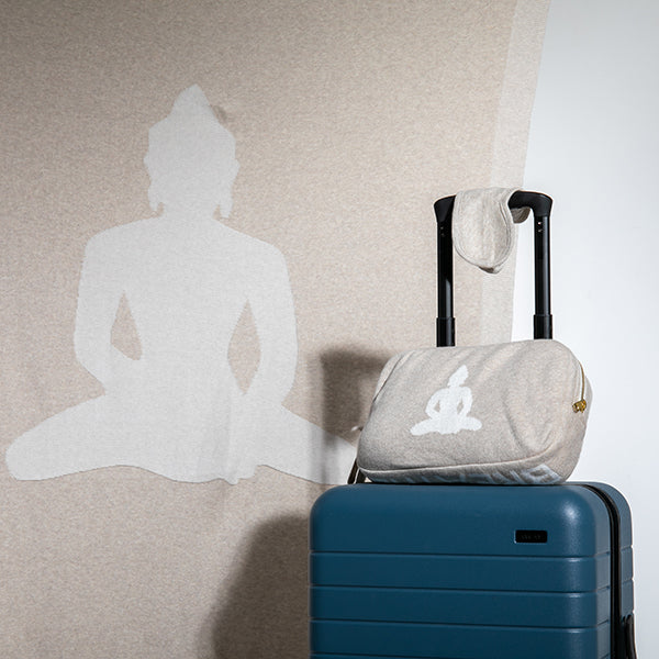Beige linen pouch with a white Buddha symbol on top of a blue suitcase next to a wall with a Buddha silhouette.