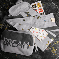 Gray hooded travel blanket, eye mask, and cotton pouch with 'Dream' printed on it on a dark granite surface.