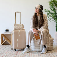 Woman sitting on a beige knot cushion with a suitcase and wearing an eye mask and a leopard print blanket.