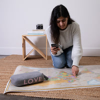 Woman using a mobile and pointing to a world map with a light gray cotton pouch labeled 'LOVE' in neon pink nearby.