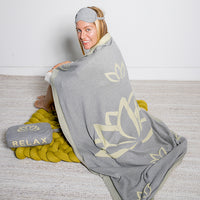 Woman wrapped in a gray blanket with lotus flower designs, sitting on a knitted yellow mat with a pouch and eye mask.