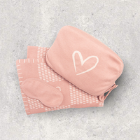 Pink checkered travel blanket, eye mask, and blanket pouch with heart design on a gray textured surface.