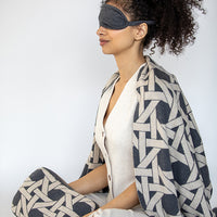 Woman wearing a patterned blanket and grey eye mask on a plain background.