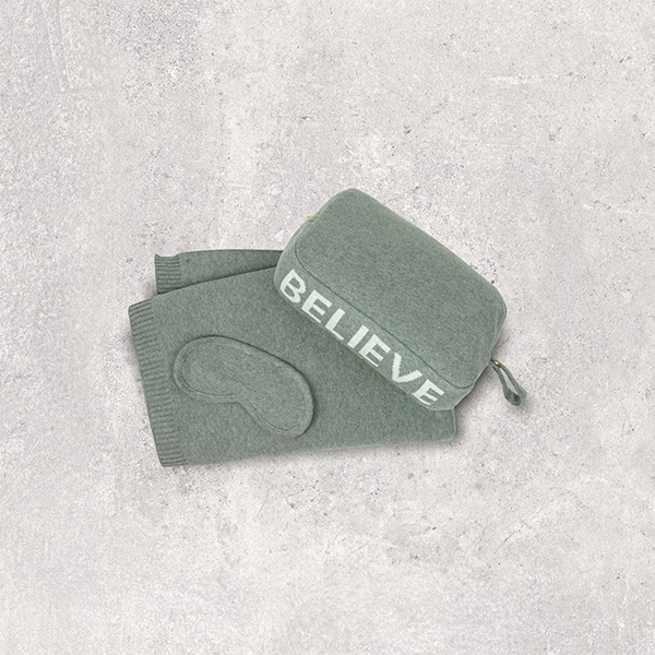 Gray pouch with 'BELIEVE' text and matching travel blanket and travel eye mask on a light gray textured background.