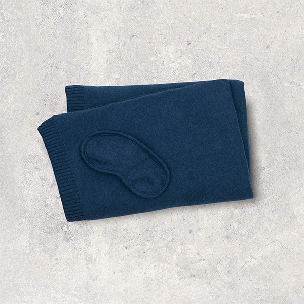 Folded cotton navy blue travel blanket with a matching eye mask on a light gray surface.