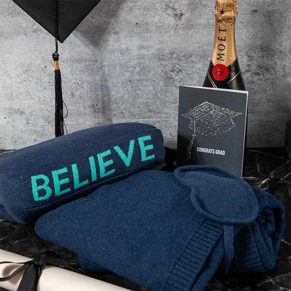 Cotton navy blue blanket with 'BELIEVE' text, bottle of Moët champagne, and graduation card on a dark surface.
