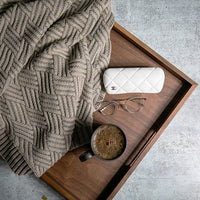Wooden tray with a chunky knit ash coloured blanket, mug, glasses, and glasses on a concrete surface.