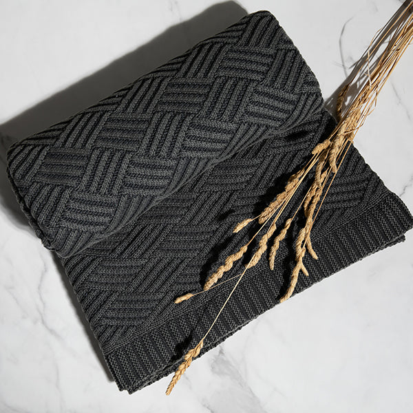 Dark gray chunky knit cotton blanket with a geometric brick pattern with wheatgrass on a light background.