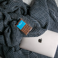 Apple laptop under a textured washed navy mel chunky knit cotton blanket with a chocolate bar on top.