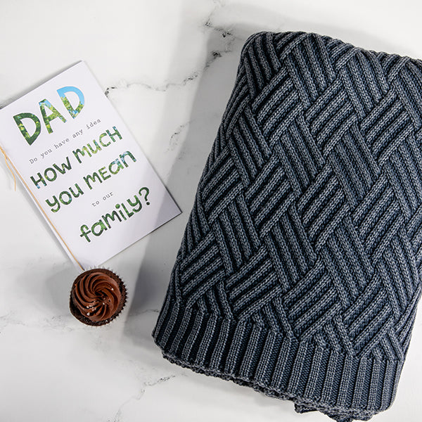 Navy mel moss-knit cotton throw blanket with a card addressed to dad and small cupcake on a marble surface.