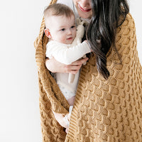 Woman holding a baby wrapped in a bronze knitted blanket against a white background.