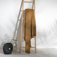 Bronze and textured knitted cotton blanket draped over a wooden ladder against a white wall next to two vases.