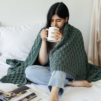 Woman sitting on a bed wrapped in a green knit throw blanket, drinking from a white mug.
