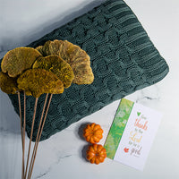 Green textured cotton blanket with curvy knit patterns place next to dried flowers and decorative pumpkins on a marble surface.