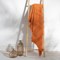 Orange textured cotton blanket draped over a wooden ladder against a white wall with two glass bottles on the floor.