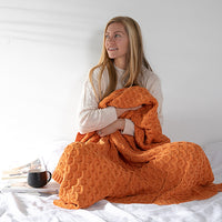Woman wrapped in an orange cotton knit blanket and sitting cross-legged on a bed with a ceramic cup.