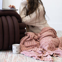 Person sitting on a textured mat with a pink knitted blanket and a mug, working on a silver laptop that is placed on a burgundy knot pillow.