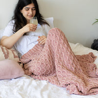 Woman sitting on a bed with a pink knitted cotton blanket, holding a drink in hand.