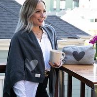Woman on a balcony wrapped with a grey Hearts Reversible blanket with a coffee mug and heart travel pouch on the table.