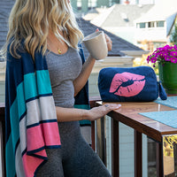 Woman holding a coffee mug on a balcony with a blue pouch featuring pink lips and a plant in the background.