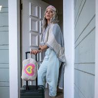 A woman wrapped in the soft grey and beige heart-design blanket, wearing the matching grey sleep mask and holding her suitcase with a compact multi heart case.