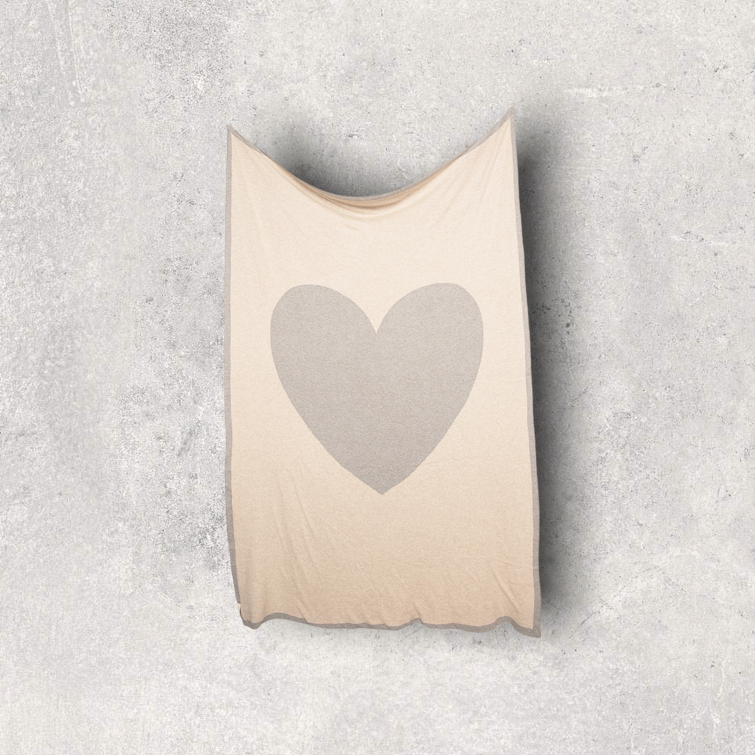 Neutral coloured cotton travel blanket with grey border heart design hanging on a wall.