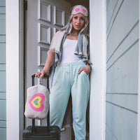 Woman wrapped in a light grey travel blanket, with a pink and grey heart eye mask leaving home with her suitcase.