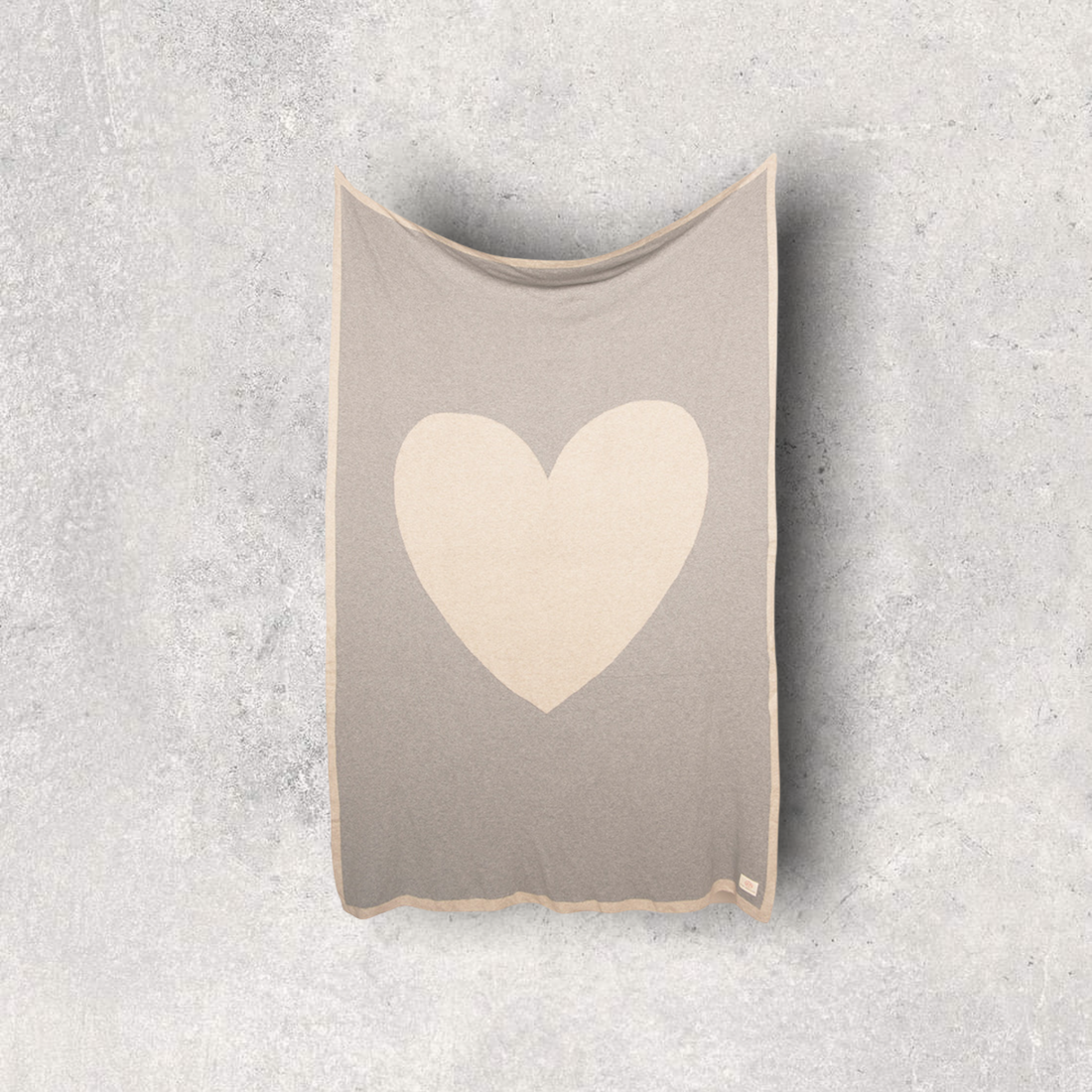 Grey cotton travel blanket with a beige heart and border hanging on a wall.