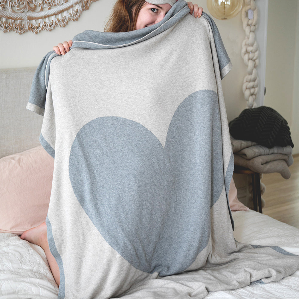 Woman holding up a luxurious beige cotton travel blanket with a grey heart and border.