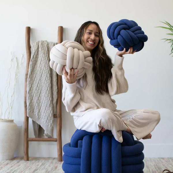 Woman holding Pink Lemonade knotted cushions while sitting on a blue knit pouf ottoman in a bright living space.