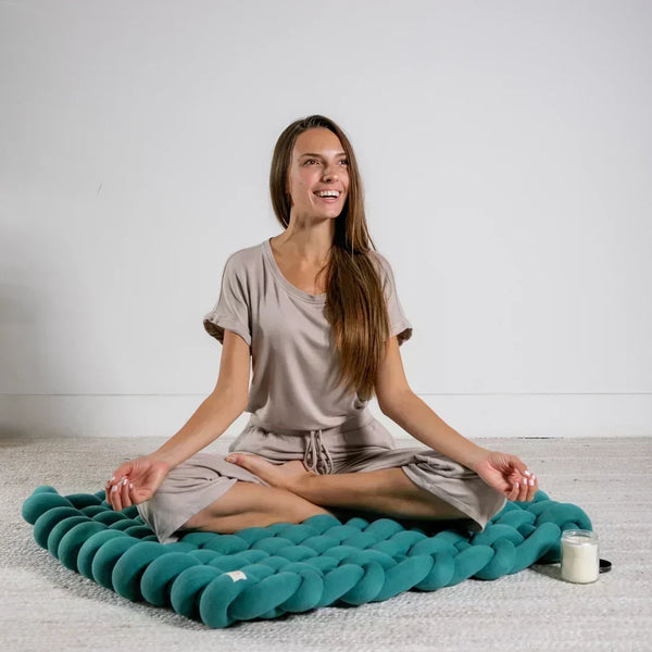 Woman meditating on a teal Pink Lemonade knotted floor cushion in a calm room.