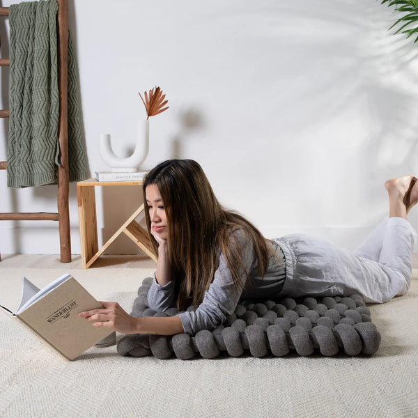 A woman reads on a gray knit floor cushion from Pink Lemonade in a bright, minimalist bedroom.
