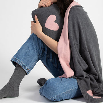 A woman wrapped in the grey & pink Hearts Reversible blanket, hugging the compact travel bag with a pink heartwhich doubles as a pillow.