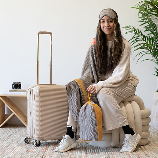 Woman sitting on a chair with a suitcase and travel bag, wearing a chevron sleep mask and holding a grey chevron cotton travel pouch