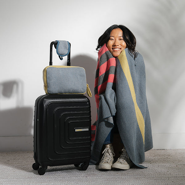 Person wrapped in a chevron design cotton blanket next to a black suitcase with a gray chevron travel pouch on a light background