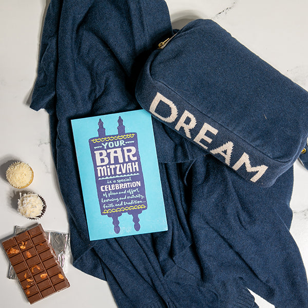 Navy blue travel blanket with the word 'dream' on it, Bar Mitzvah card, chocolate, and cupcakes on a light background.