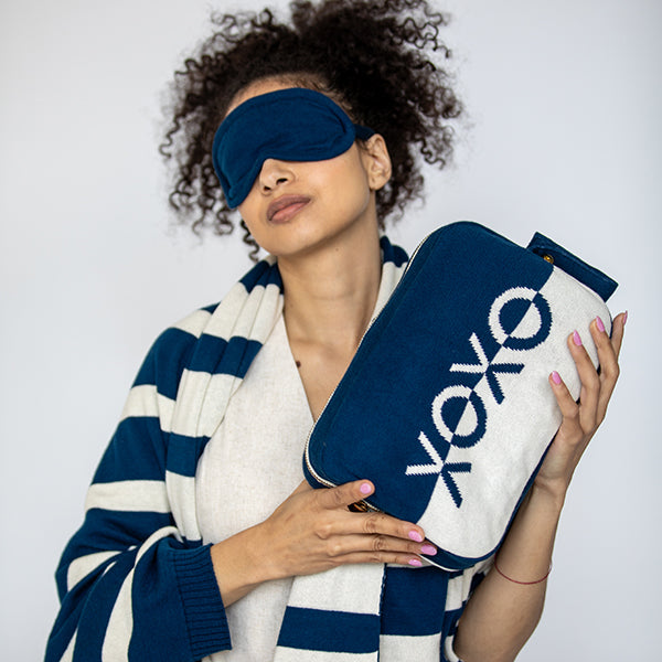 Woman wearing a blue sleep mask and holding a XOXO branded cotton pouch against a white background
