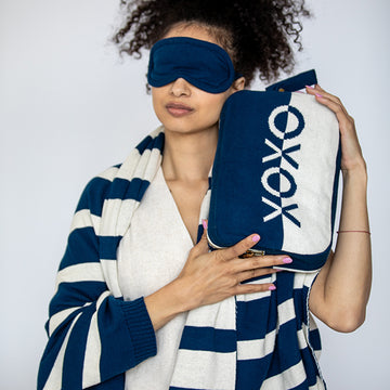 Woman with a blue eye mask holding a blue and white striped blanket with a matching case with 'XOXO' text against a plain background.