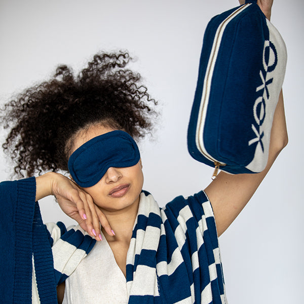 Woman wearing a blue cotton sleep mask and holding a navy blue and white bag.