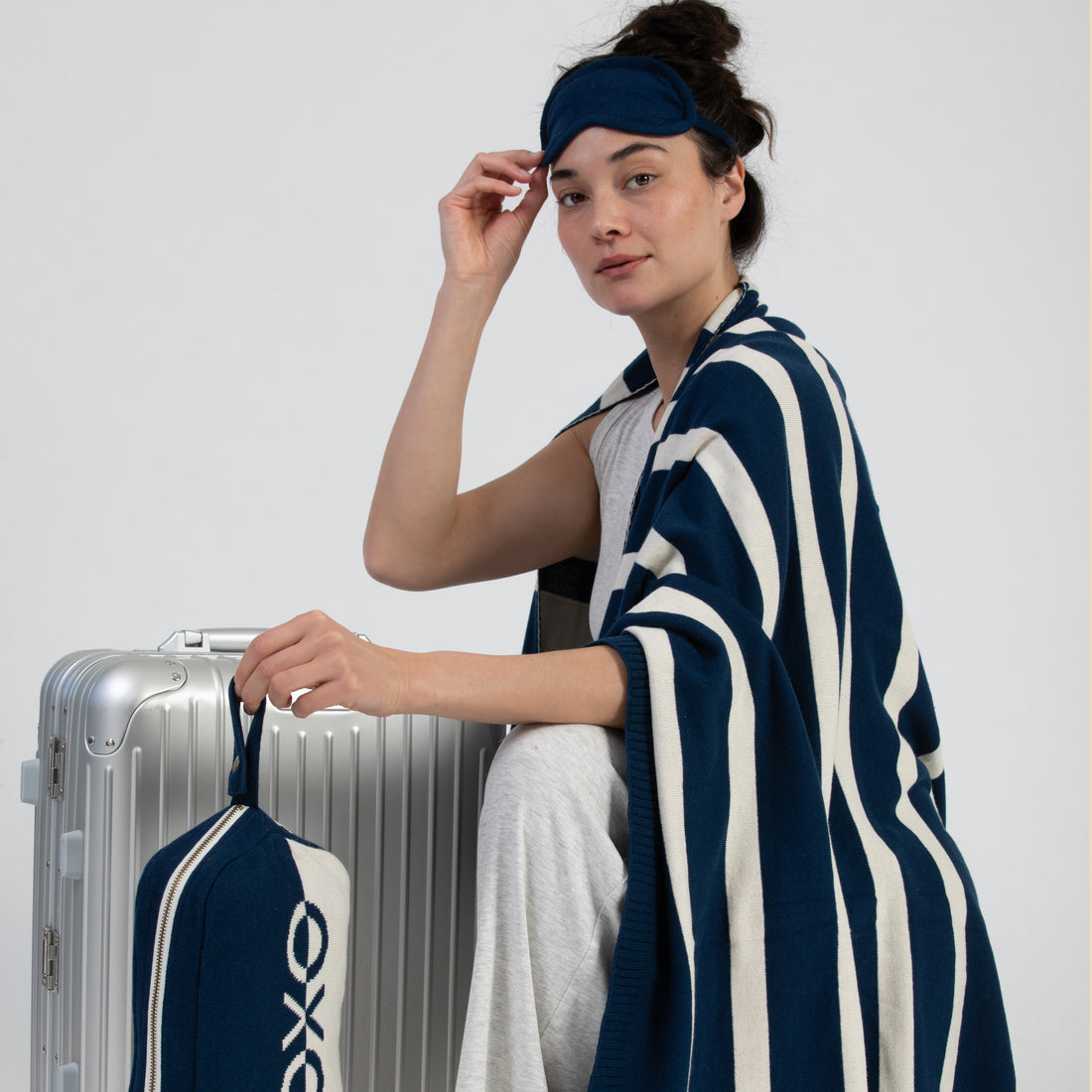 Woman wrapped in a striped navy and white blanket holding a silver suitcase with a blue eye mask on her head against a white background