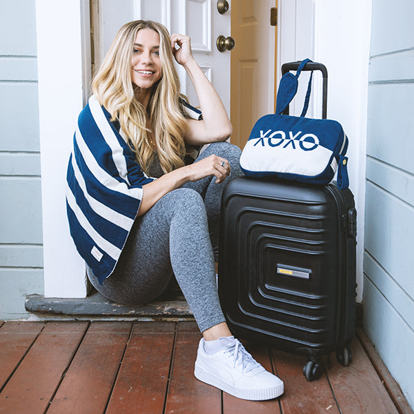 Woman sitting on a step with a black suitcase and a blue and white travel pouch with 'XOXO' text.