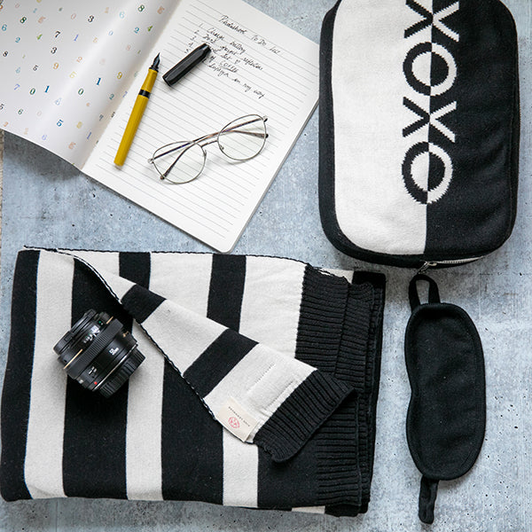Black and white striped pouch, camera lens, eyeglasses, notebook, pen, and black sleep mask on a gray surface with 'XOXO' pouch.