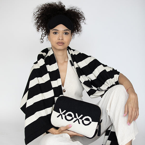 Woman holding a black and white pouch with 'XOXO' text, wearing a black and white striped blanket and black eye mask.