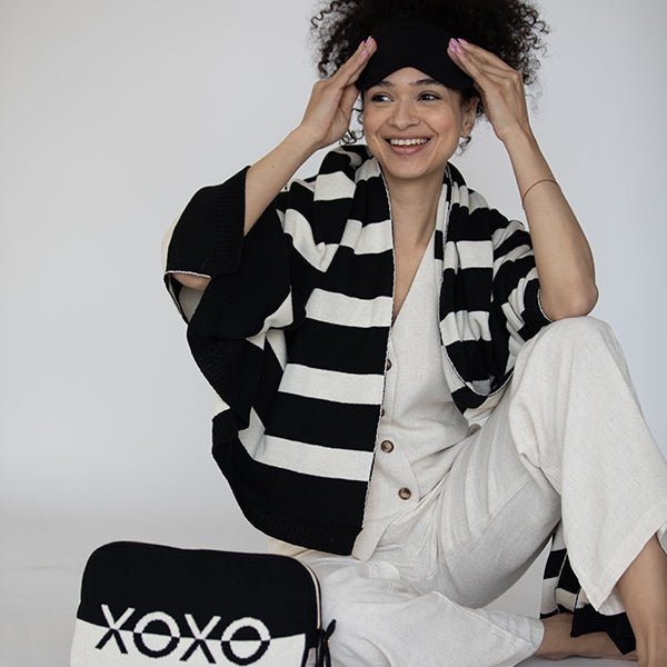Woman in a white outfit with a black and white striped blanket around her, sitting next to a pouch with 'XOXO' branding.