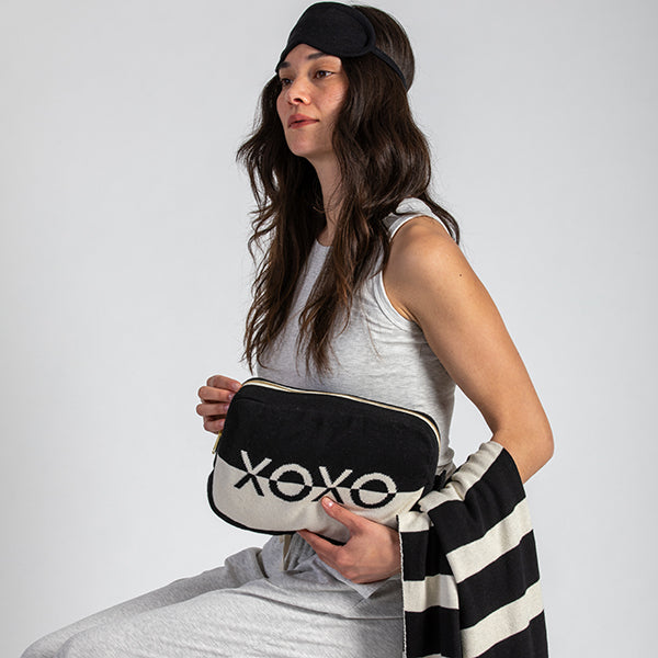 Woman holding a black and white pouch with 'XOXO' text, wearing a sleep mask and striped blanket.