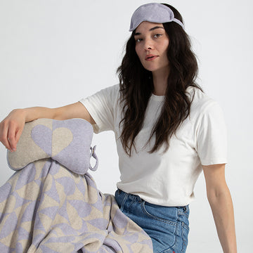 Woman wearing a lavender sleep mask and holding a blanket pouch with a heart and wearing a grey blanket with geometric patterns on a plain background