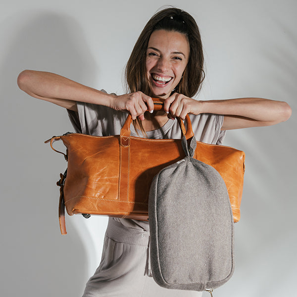 Woman smiling and holding a brown leather bag and a gray fabric pouch against a plain background.