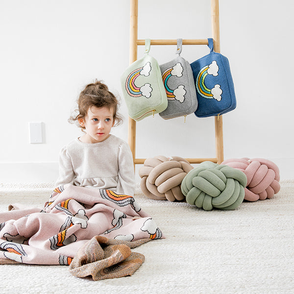 Child sitting on a rug and wrapped in a soft pink cotton blanket with rainbow patterns next to three knotted cushions and blanket pouches. 